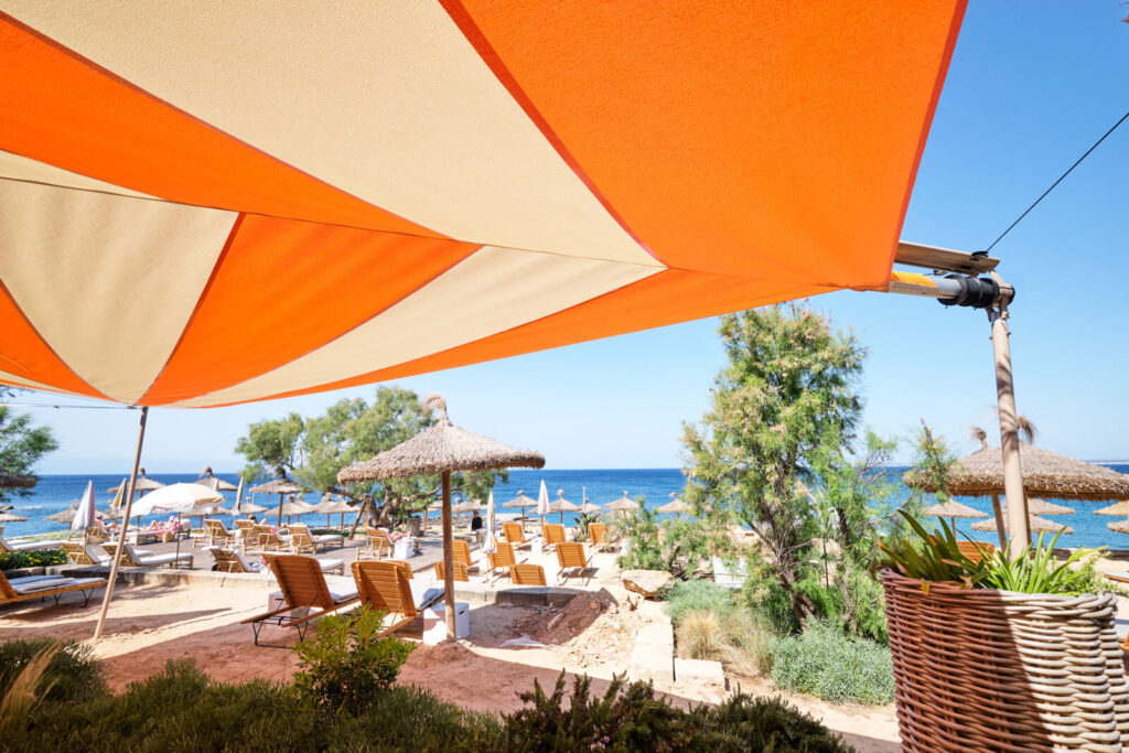 Busy lunch service on Mallorcan seafront restaurant terrace with SunSquare automatic shade deployed overhead, guests and wait staff in frame